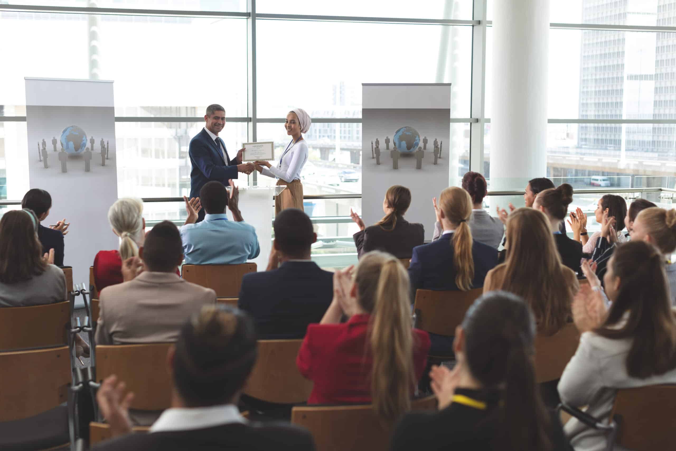 Front view of mixed race businesswoman receiving award from mixed race businessman in front of business professionals applauding at business seminar in office building