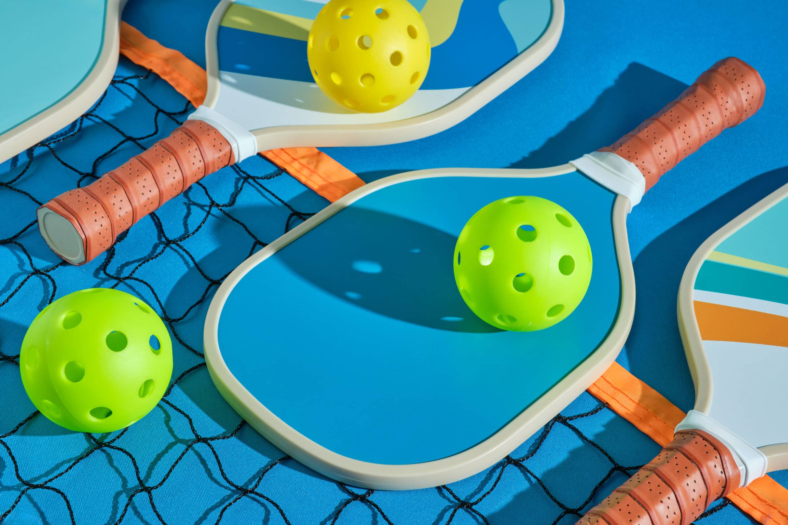 pickleball paddles with balls on top of a net on a blue background illuminated with hard light