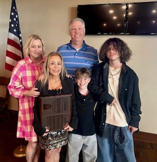 A family of five posing indoors with an American flag in the background; the woman in pink plaid holds a large award plaque while the others smile.