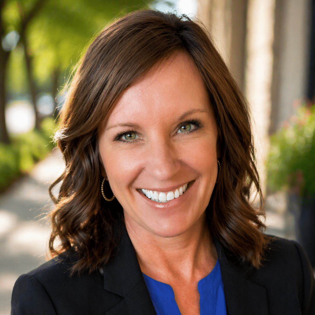 Smiling woman with brown hair in a blue blouse and black blazer, outdoors on a sunny day.