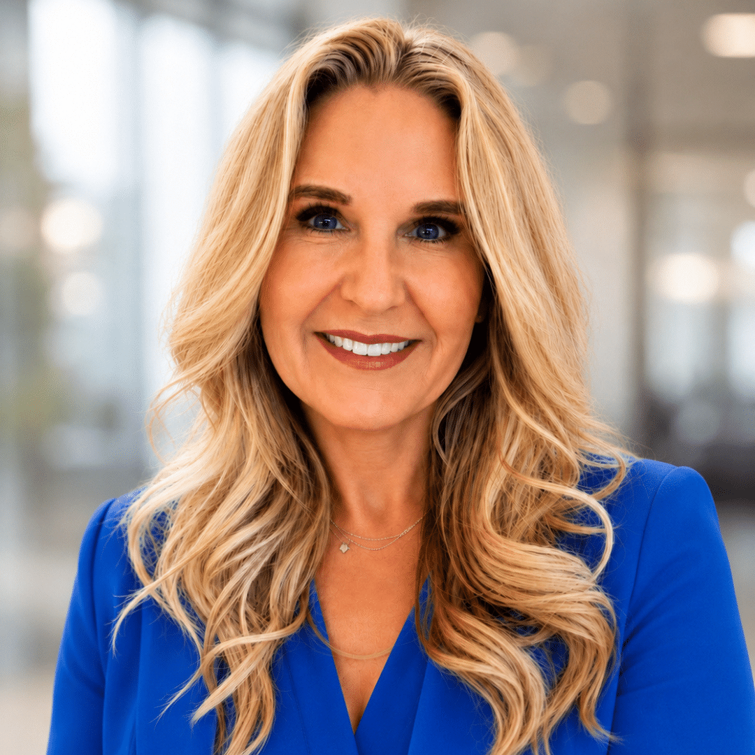 Professional portrait of a smiling blonde woman in a royal blue blazer in a modern office setting.