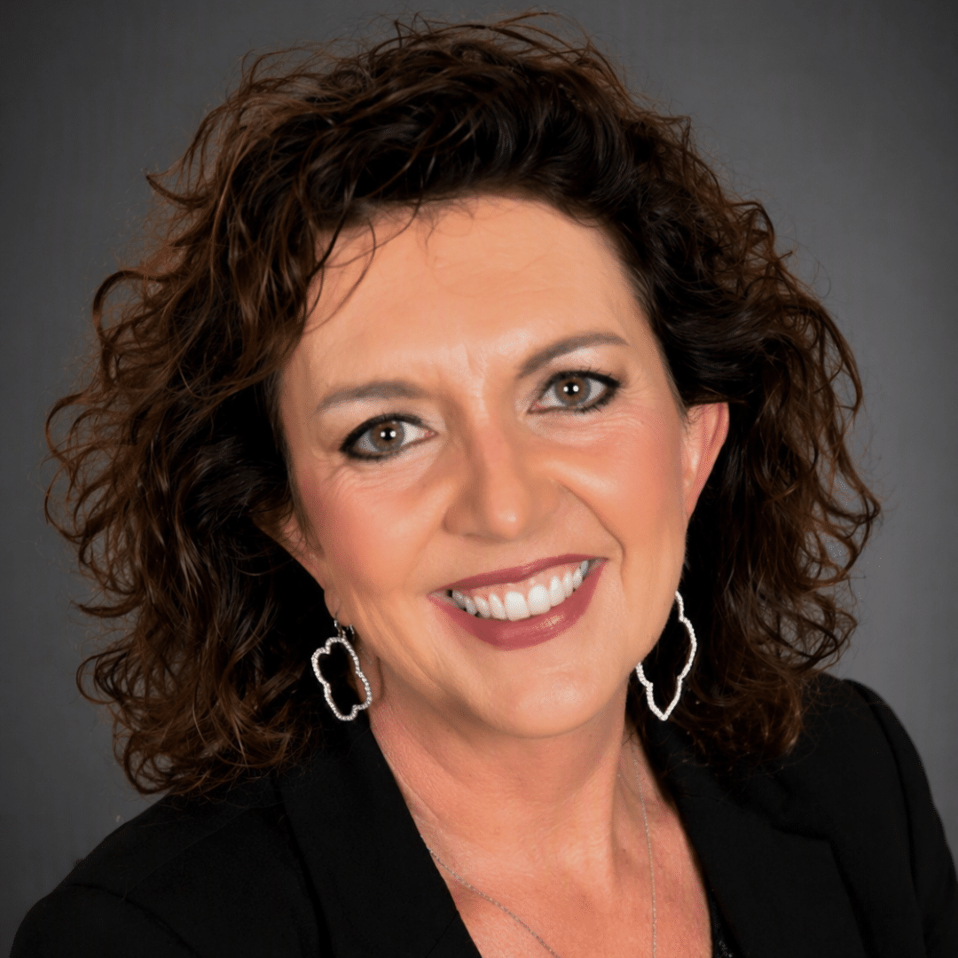 Smiling woman with curly brown hair in a black blazer, silver hoop earrings, against a gray backdrop.