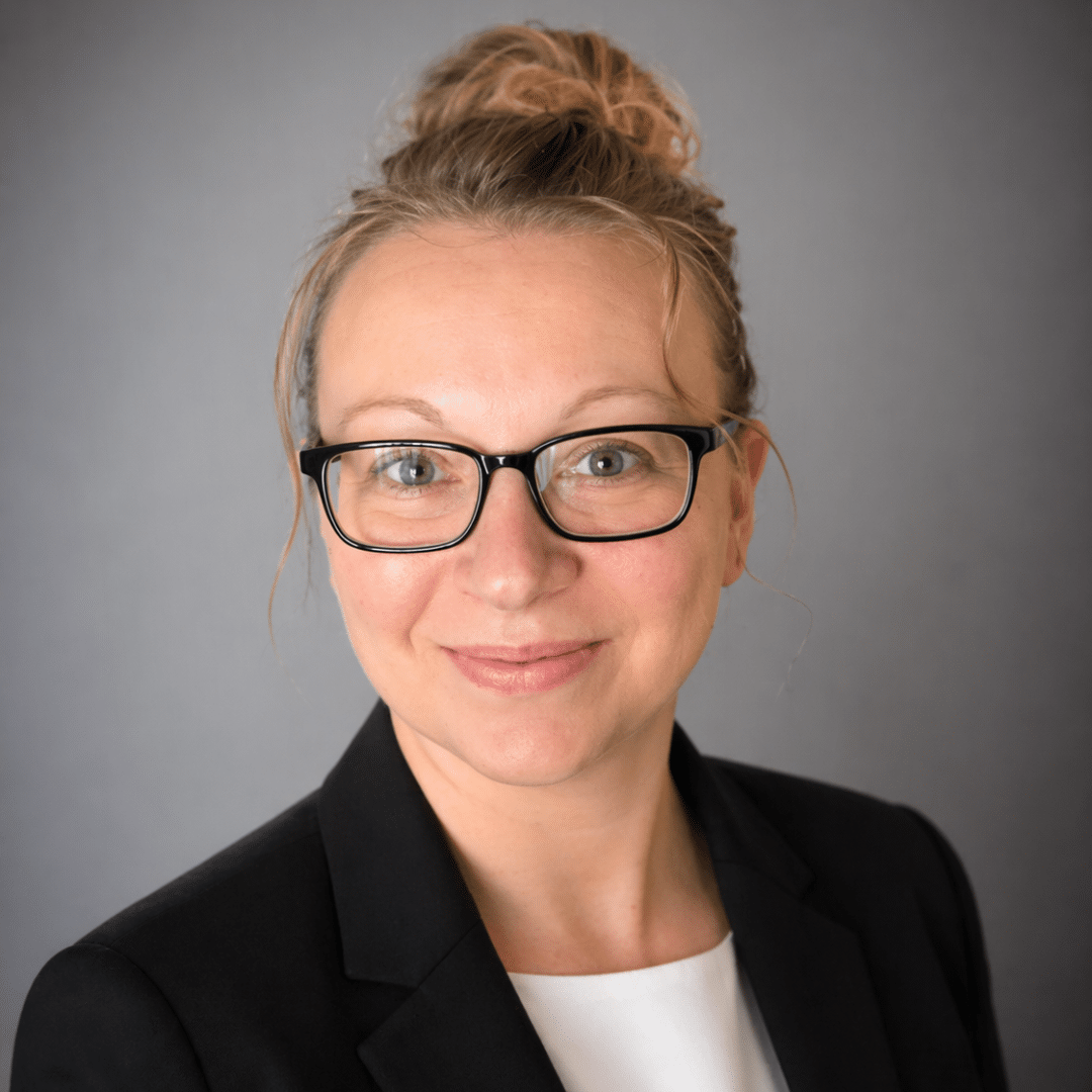 Professional headshot of a smiling woman with black glasses, in a black blazer against a gray background.