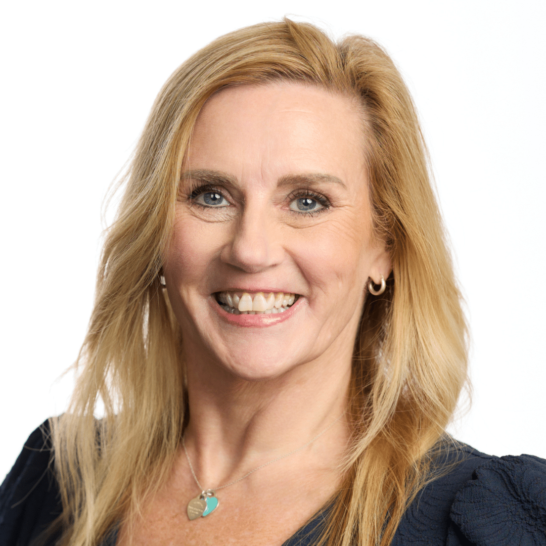 Smiling blonde woman in a navy top with a heart pendant necklace, against a white background (head-and-shoulders portrait).